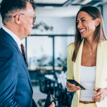 Two businesspeople stand indoors, smiling and talking. The man in a blue suit holds a wallet, while the woman in a yellow suit holds a phone. Office furniture and large windows are in the background.