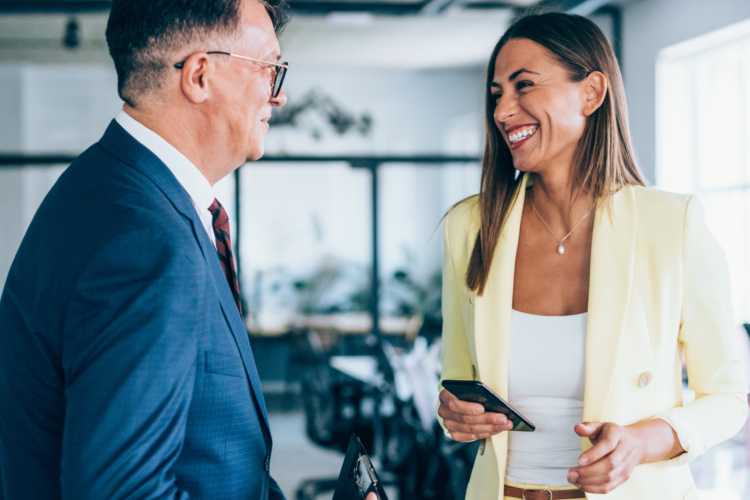Two businesspeople stand indoors, smiling and talking. The man in a blue suit holds a wallet, while the woman in a yellow suit holds a phone. Office furniture and large windows are in the background.