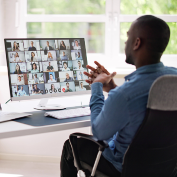 Man in blue shirt sits at desk participating in a large video conference with multiple people visible on a computer monitor in a bright office setting.