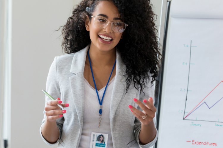 Woman in business attire presenting data on a flip chart showing sales and expenditures lines, standing in a bright office, holding a marker, and wearing a conference badge.