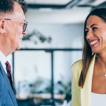 Two people in business attire stand indoors, facing each other and smiling, in a well-lit, modern office setting.