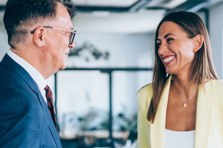 Two people in business attire stand indoors, facing each other and smiling, in a well-lit, modern office setting.