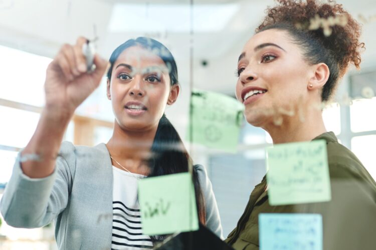Two women collaborate, writing on a transparent board covered with notes and sticky notes in a modern office setting.