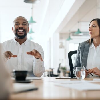 Two professionals sit at a conference table having a discussion in a modern office setting, with documents, a glass of water, and a coffee cup on the table.