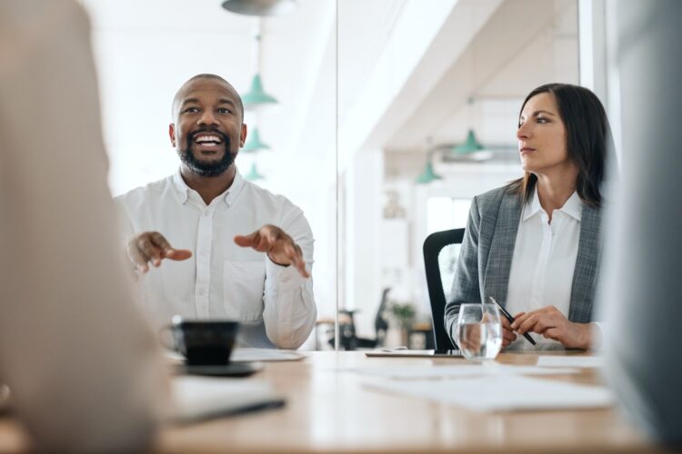 Two professionals sit at a conference table having a discussion in a modern office setting, with documents, a glass of water, and a coffee cup on the table.