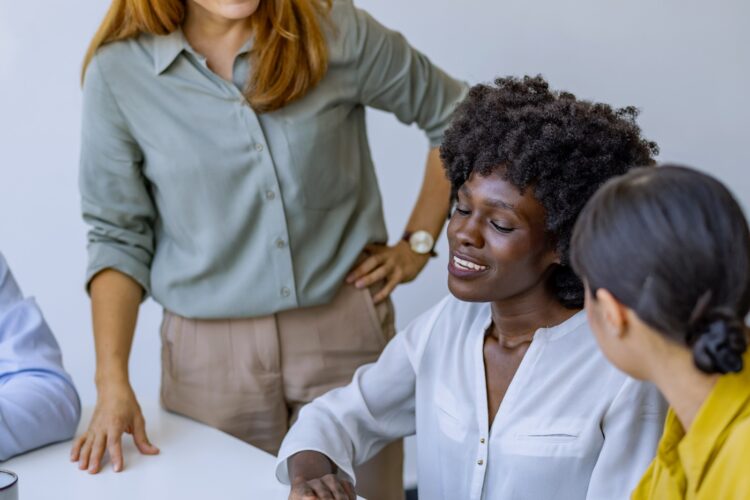Three women discuss architectural plans at a table, with one woman pointing at a blueprint while the others listen and engage.