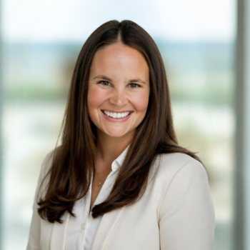 Woman with long brown hair wearing a white blazer smiles at the camera, standing indoors with blurred windows in the background.