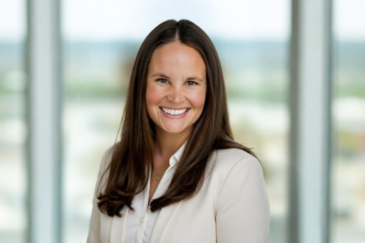Woman with long brown hair wearing a white blazer smiles at the camera, standing indoors with blurred windows in the background.