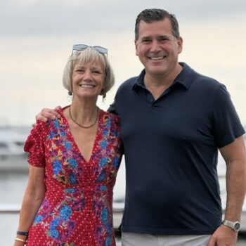 A woman in a red floral dress and a man in a navy polo shirt pose together outdoors near a marina.