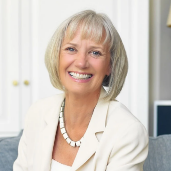A smiling woman with blonde hair, wearing a white blazer and a statement necklace.