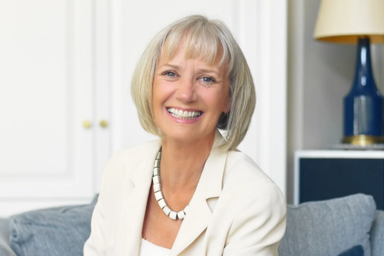 A smiling woman with blonde hair, wearing a white blazer and a statement necklace.
